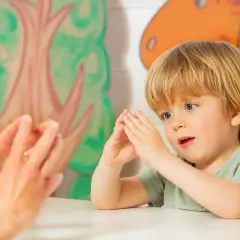 Ein Junge mit blonden Haaren und blauen Augen macht Fingerspiele im Kindergarten. Er schaut gespannt zu, wie die Hände einer Erwachsenen vor ihm tanzen.