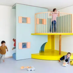 Kinder erkunden eine bunte Lernwerkstatt Kita. Ein Junge rennt, ein anderer versteckt sich, während ein Mädchen oben auf dem Spielturm steht.