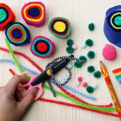 Bunte Rollen aus Papier, Pfeifenreiniger, Pompons und eine Schere laden zum Basteln ein. Ein Regenbogen-Holzstück und ein bunter Bleistift liegen bereit.
