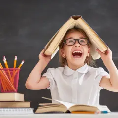 Ein fröhliches Mädchen mit Brille hält ein Buch über dem Kopf, als wäre es ein Dach. Buntstifte und Äpfel liegen auf dem Tisch. Ein Bild, das die Freude an Projekten im Kindergarten einfängt.