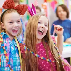 Zwei lachende Mädchen mit bunten Schleifen und Krönchen feiern ausgelassen Fasching in der Kita.