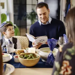 Familie feiert Schabbat. Vater liest aus einem Buch mit Davidstern. Der Sohn, der gerade das Judentum in der Grundschule lernt, hört aufmerksam zu.