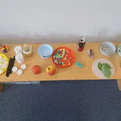 Tisch mit Materialien für Experimente: Würfel, Kerzen, Äpfel, bunte Perlen, Steine, Büroklammern, Knöpfe, Blätter und Münzen. Kinder können hier spielerisch lernen.