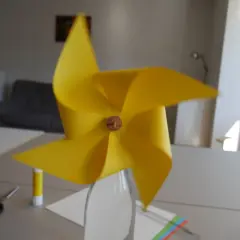 Ein gelbes Windrad, das man leicht selber bauen kann, steht in einer Glasflasche. Schere und Klebestift liegen bereit.