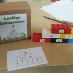Bunte Legosteine bilden eine Zahlenmauer mit aufgeschriebenen Zahlen. Daneben eine Pappbox mit gleicher Zahlenmauer.