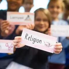 Kinder der Grundschule halten Schilder hoch, eines davon mit "Religion" beschriftet. Sie lernen über verschiedene Fächer, vielleicht im Rahmen der Fastenzeit.