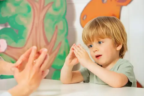 Ein Junge mit blonden Haaren und blauen Augen macht Fingerspiele im Kindergarten. Er schaut gespannt zu, wie die Hände einer Erwachsenen vor ihm tanzen.