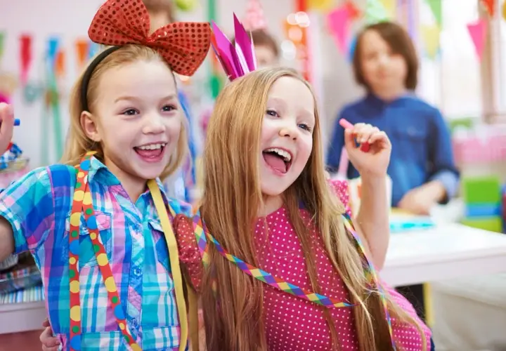 Zwei lachende Mädchen mit bunten Schleifen und Krönchen feiern ausgelassen Fasching in der Kita.