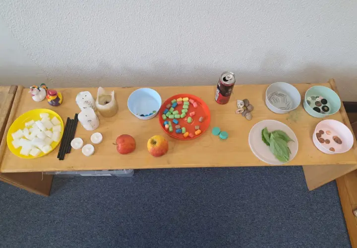 Tisch mit Materialien für Experimente: Würfel, Kerzen, Äpfel, bunte Perlen, Steine, Büroklammern, Knöpfe, Blätter und Münzen. Kinder können hier spielerisch lernen.