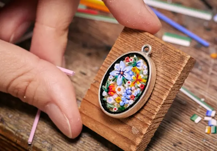 Ein zartes Blumen-Mosaik, einfach und detailreich, wird von Fingern gehalten. Kleine Glasstäbchen liegen bereit.