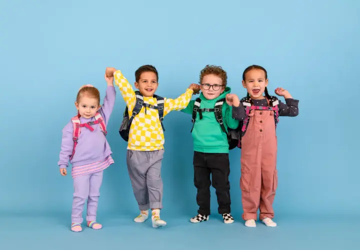 Vier fröhliche Kinder mit Rucksäcken, bereit für die Schule. Ein kleiner Junge mit kariertem Pullover scheint begeistert, vielleicht ist er ein **kind vorzeitig einschulen**?