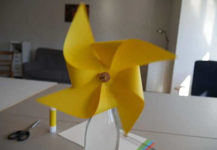 Ein gelbes Windrad, das man leicht selber bauen kann, steht in einer Glasflasche. Schere und Klebestift liegen bereit.