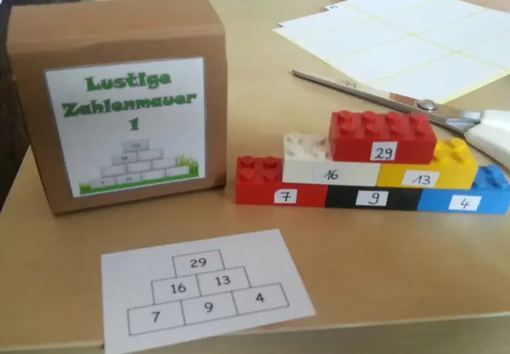 Bunte Legosteine bilden eine Zahlenmauer mit aufgeschriebenen Zahlen. Daneben eine Pappbox mit gleicher Zahlenmauer.