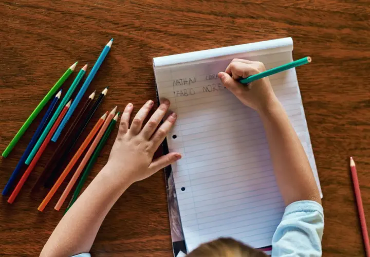 Kind schreibt Namen auf Papier, Teil der schuleingangsuntersuchung was wird gemacht. Buntstifte liegen bereit.