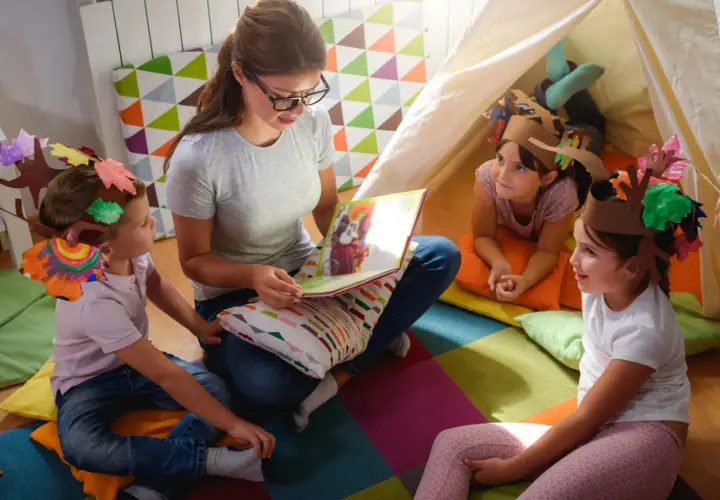 Lehrerin liest Kindern im Kindergarten eine Geschichte vor. Die Kinder tragen selbstgebastelte Baumkronen, ein Beispiel für Nachhaltigkeit im Kindergarten.