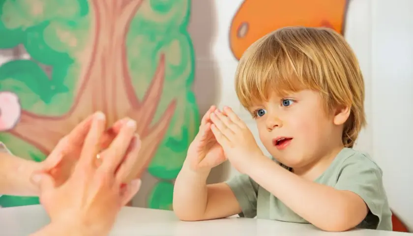 Ein Junge mit blonden Haaren und blauen Augen macht Fingerspiele im Kindergarten. Er schaut gespannt zu, wie die Hände einer Erwachsenen vor ihm tanzen.