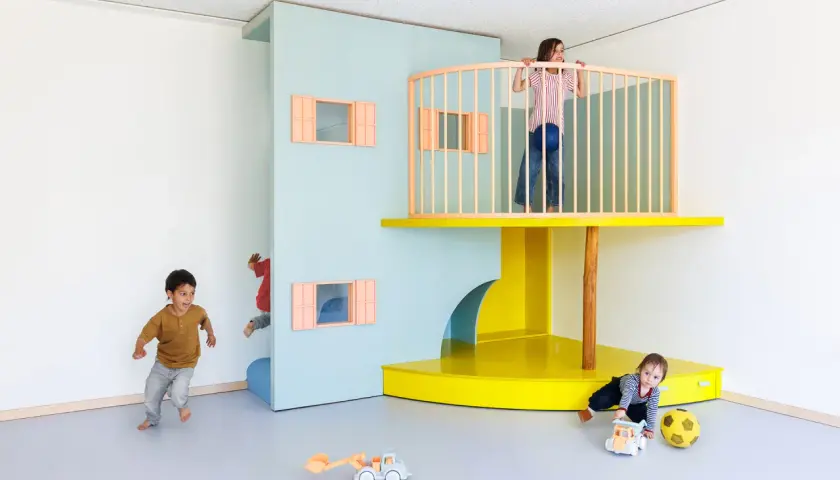 Kinder erkunden eine bunte Lernwerkstatt Kita. Ein Junge rennt, ein anderer versteckt sich, während ein Mädchen oben auf dem Spielturm steht.