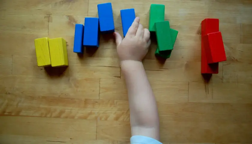 Ein Kinderarm greift nach bunten Holzklötzen. So lernen Kinder spielerisch Farben kennen: Gelb, Blau, Grün und Rot.