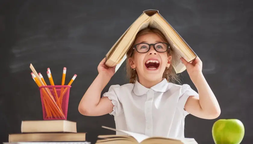 Ein fröhliches Mädchen mit Brille hält ein Buch über dem Kopf, als wäre es ein Dach. Buntstifte und Äpfel liegen auf dem Tisch. Ein Bild, das die Freude an Projekten im Kindergarten einfängt.