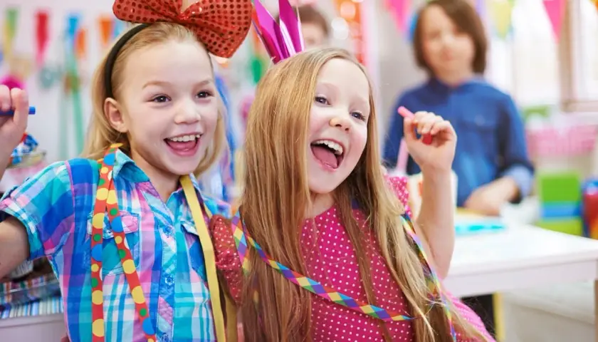 Zwei lachende Mädchen mit bunten Schleifen und Krönchen feiern ausgelassen Fasching in der Kita.