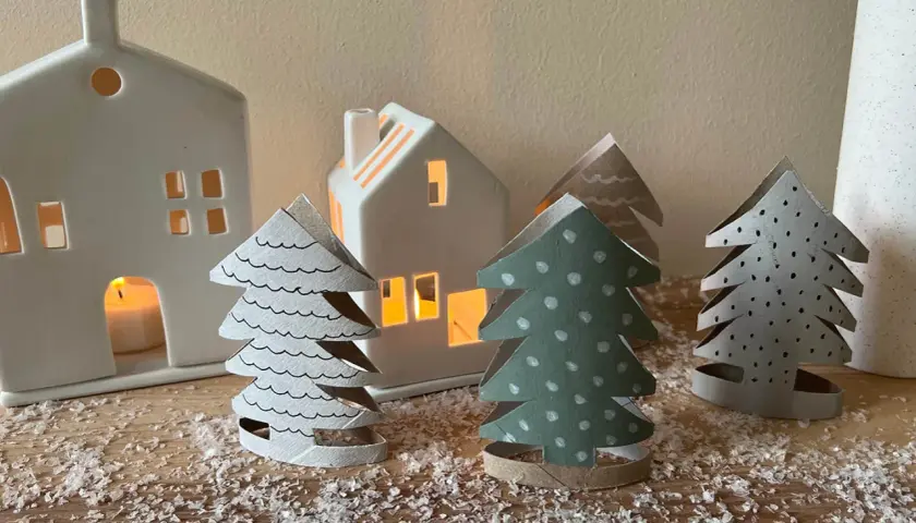 Kleine Weihnachtsbäume aus Papier, die Kinder basteln könnten, stehen vor leuchtenden Häusern.