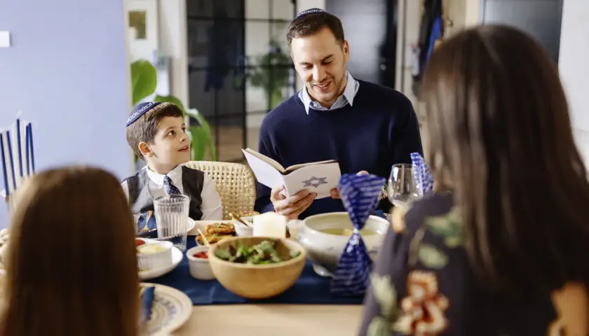 Familie feiert Schabbat. Vater liest aus einem Buch mit Davidstern. Der Sohn, der gerade das Judentum in der Grundschule lernt, hört aufmerksam zu.