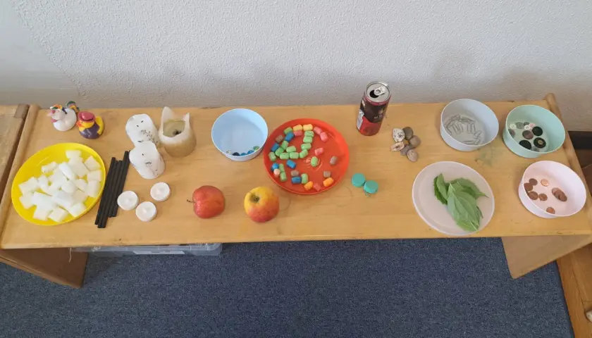 Tisch mit Materialien für Experimente: Würfel, Kerzen, Äpfel, bunte Perlen, Steine, Büroklammern, Knöpfe, Blätter und Münzen. Kinder können hier spielerisch lernen.