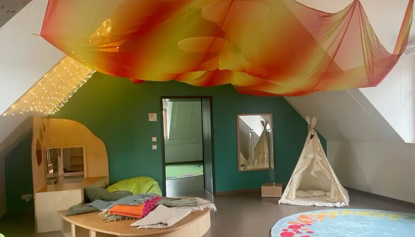 Ein gemütlicher Snoezelraum Kita mit bunten Decken, einem Spielhaus, einer Tipizelt und Lichterketten.