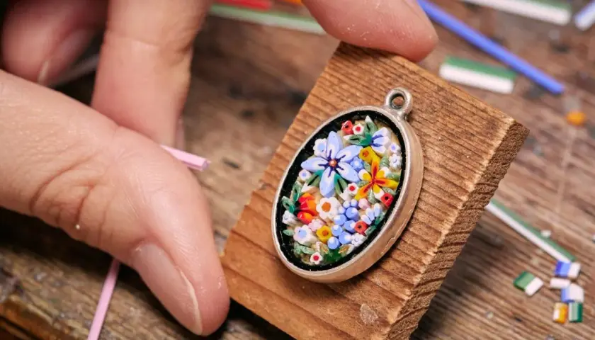 Ein zartes Blumen-Mosaik, einfach und detailreich, wird von Fingern gehalten. Kleine Glasstäbchen liegen bereit.