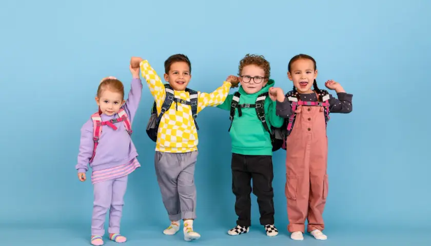 Vier fröhliche Kinder mit Rucksäcken, bereit für die Schule. Ein kleiner Junge mit kariertem Pullover scheint begeistert, vielleicht ist er ein **kind vorzeitig einschulen**?