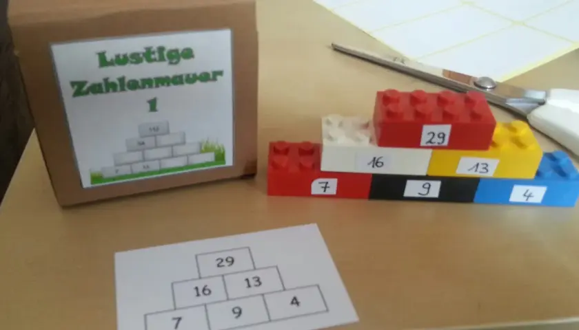 Bunte Legosteine bilden eine Zahlenmauer mit aufgeschriebenen Zahlen. Daneben eine Pappbox mit gleicher Zahlenmauer.