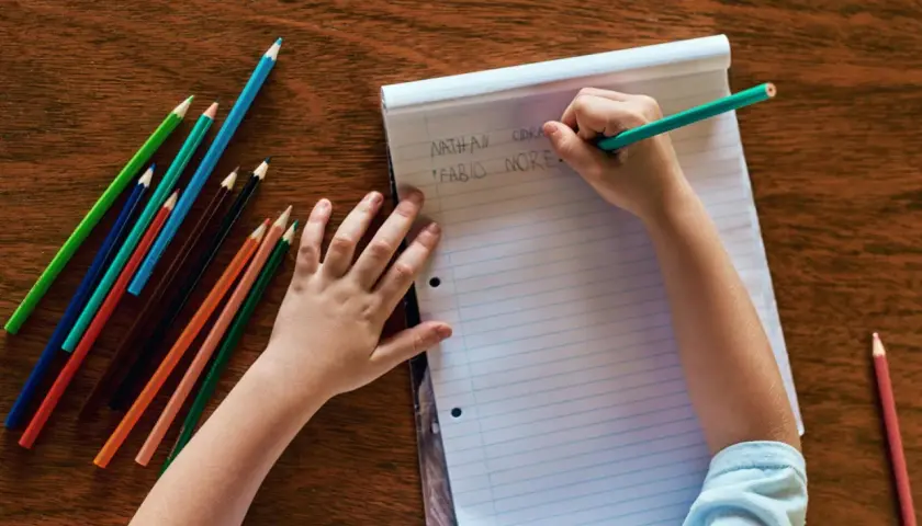 Kind schreibt Namen auf Papier, Teil der schuleingangsuntersuchung was wird gemacht. Buntstifte liegen bereit.