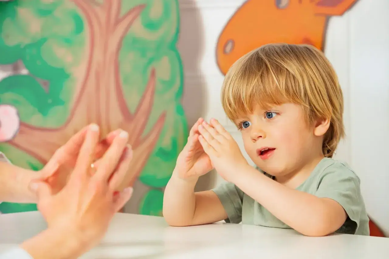 Ein Junge mit blonden Haaren und blauen Augen macht Fingerspiele im Kindergarten. Er schaut gespannt zu, wie die Hände einer Erwachsenen vor ihm tanzen.