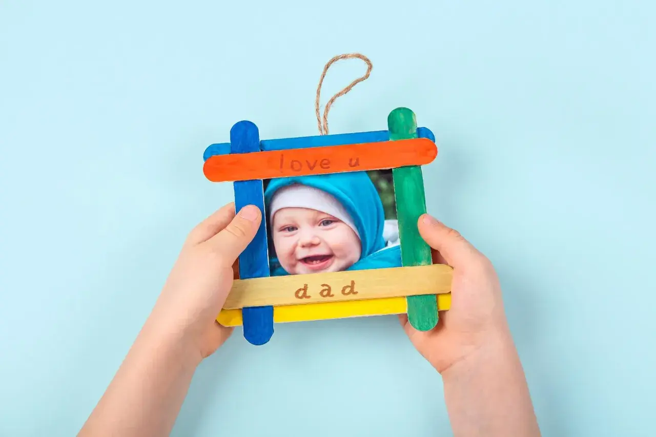 Ein süßes Vatertagsgeschenk basteln: Ein selbstgemachter Bilderrahmen aus bunten Eisstielen mit einem Foto eines lächelnden Babys und der Aufschrift "love u dad".