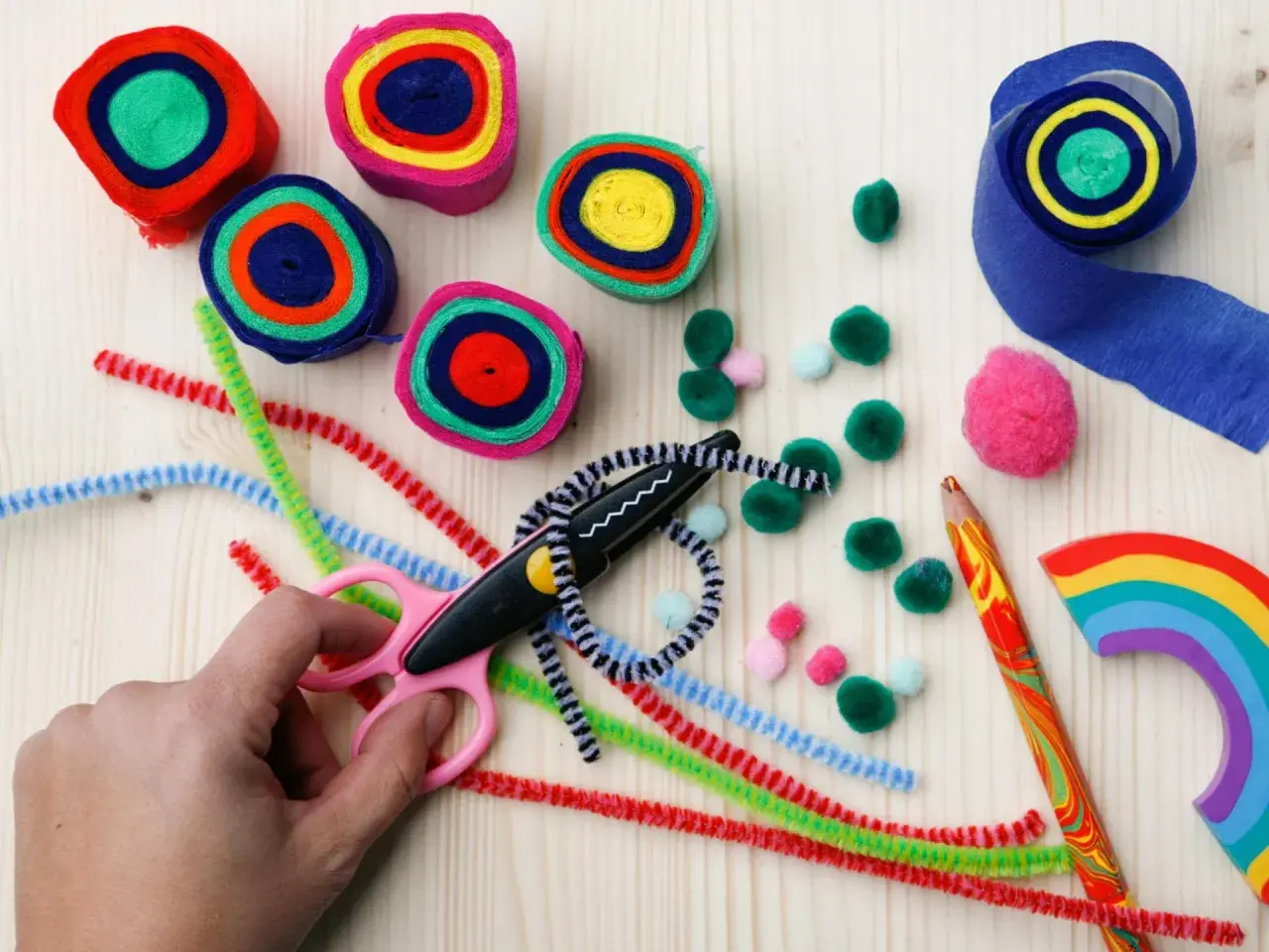 Bunte Rollen aus Papier, Pfeifenreiniger, Pompons und eine Schere laden zum Basteln ein. Ein Regenbogen-Holzstück und ein bunter Bleistift liegen bereit.
