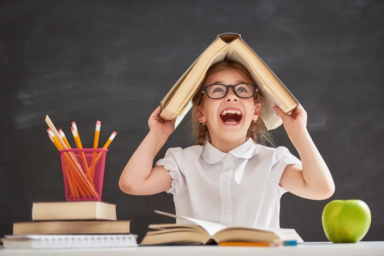 Ein fröhliches Mädchen mit Brille hält ein Buch über dem Kopf, als wäre es ein Dach. Buntstifte und Äpfel liegen auf dem Tisch. Ein Bild, das die Freude an Projekten im Kindergarten einfängt.
