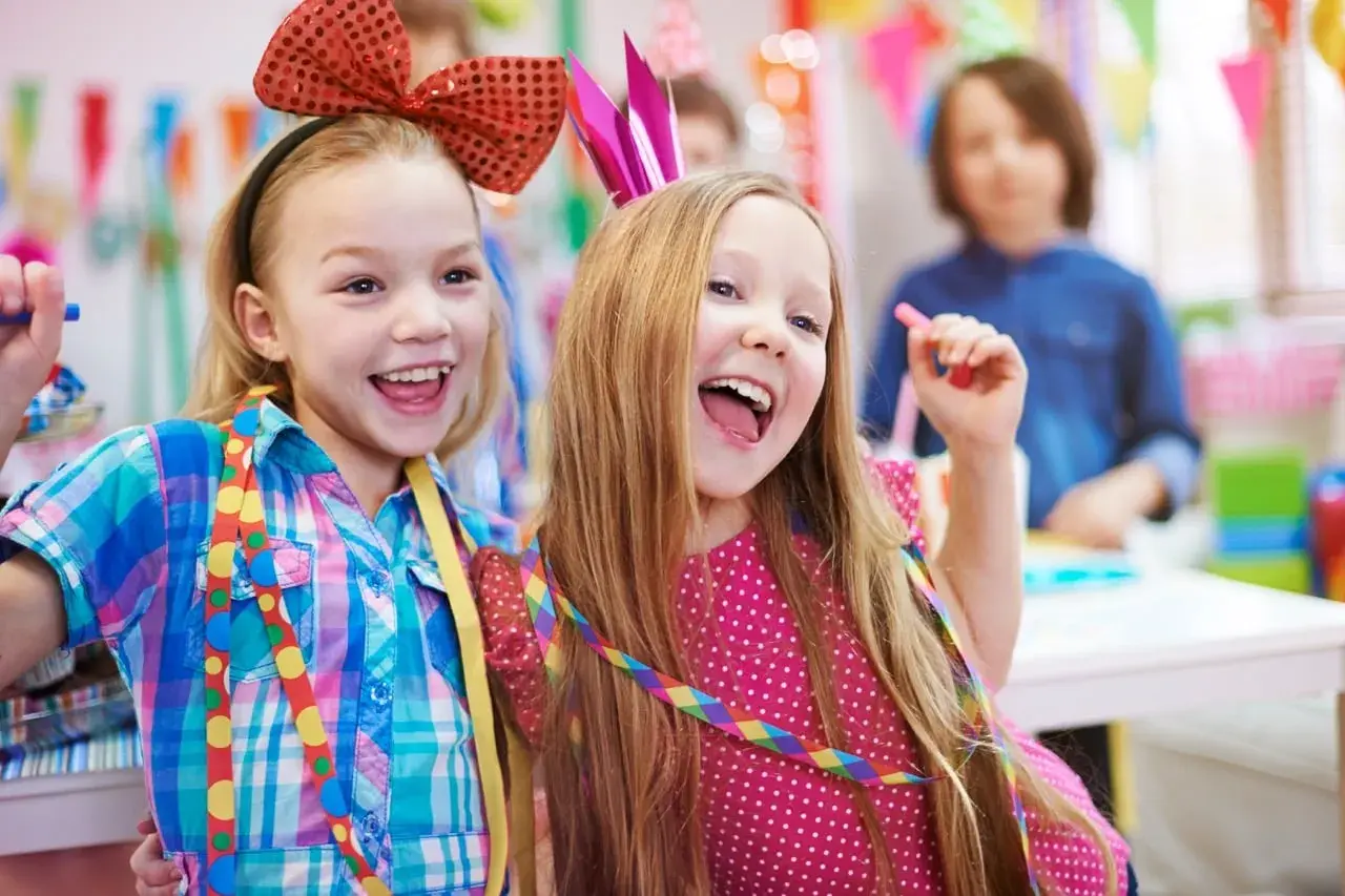 Zwei lachende Mädchen mit bunten Schleifen und Krönchen feiern ausgelassen Fasching in der Kita.