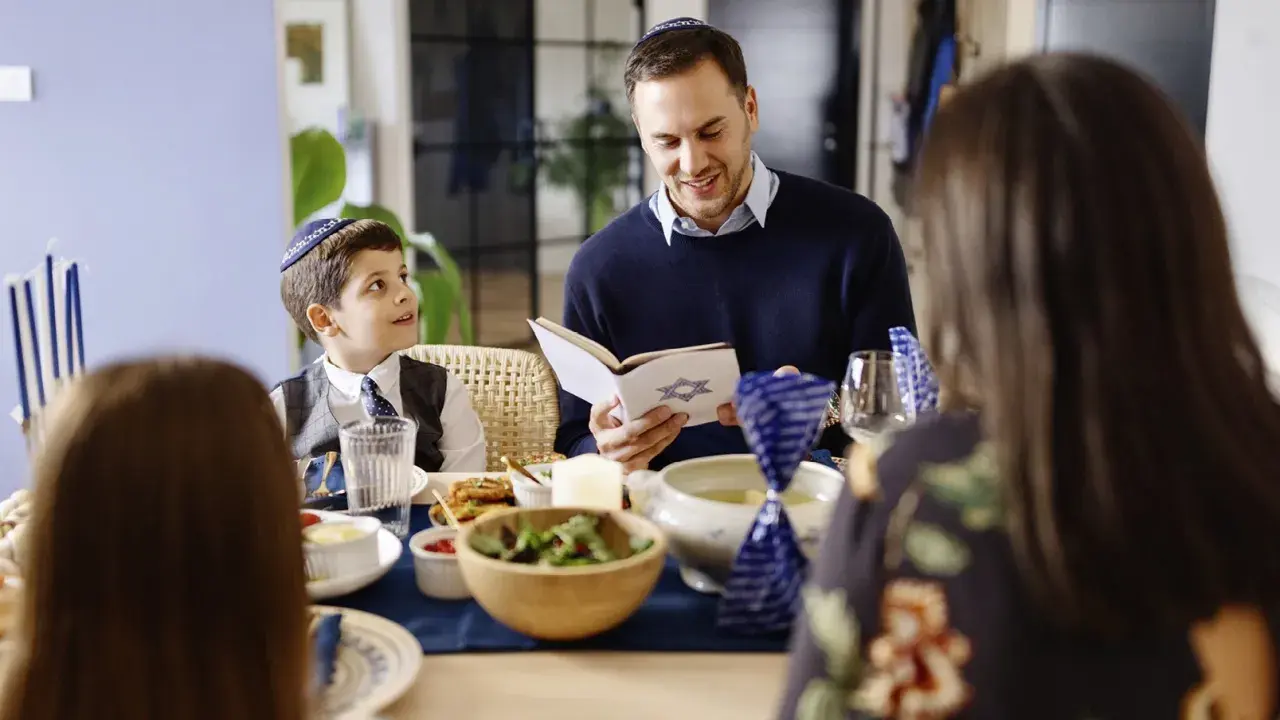 Familie feiert Schabbat. Vater liest aus einem Buch mit Davidstern. Der Sohn, der gerade das Judentum in der Grundschule lernt, hört aufmerksam zu.