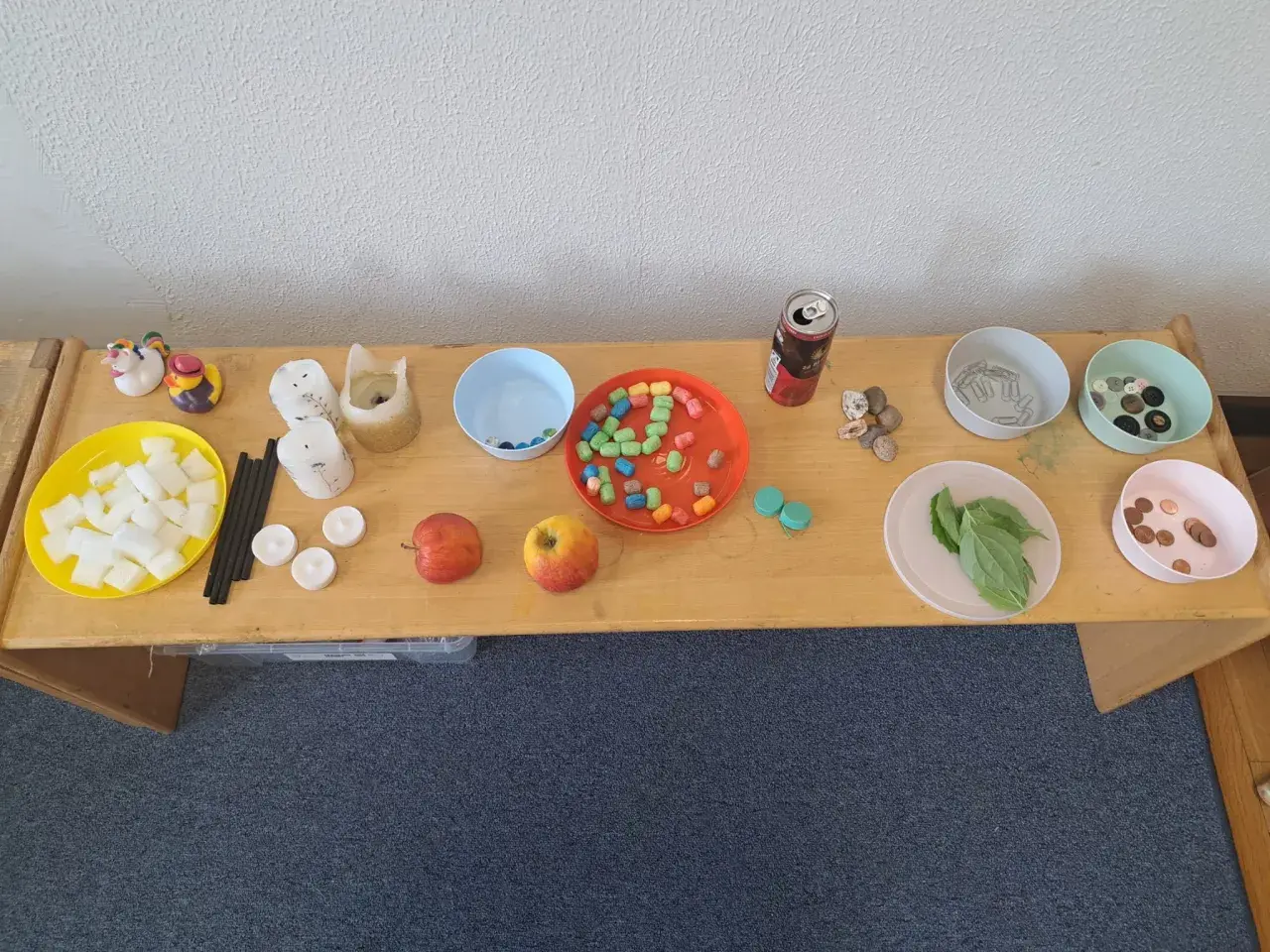 Tisch mit Materialien für Experimente: Würfel, Kerzen, Äpfel, bunte Perlen, Steine, Büroklammern, Knöpfe, Blätter und Münzen. Kinder können hier spielerisch lernen.