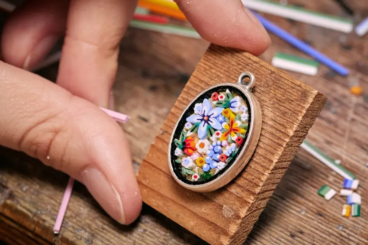Ein zartes Blumen-Mosaik, einfach und detailreich, wird von Fingern gehalten. Kleine Glasstäbchen liegen bereit.