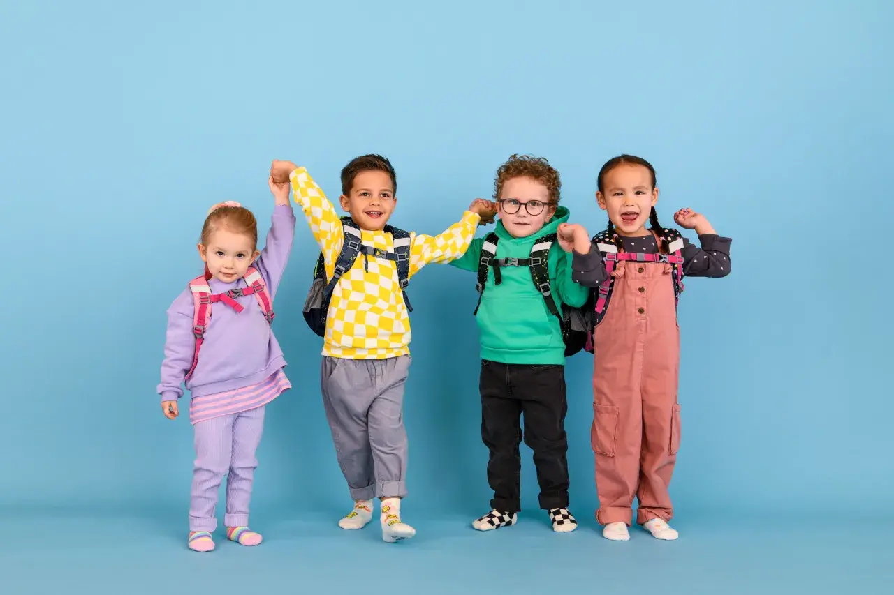 Vier fröhliche Kinder mit Rucksäcken, bereit für die Schule. Ein kleiner Junge mit kariertem Pullover scheint begeistert, vielleicht ist er ein **kind vorzeitig einschulen**?
