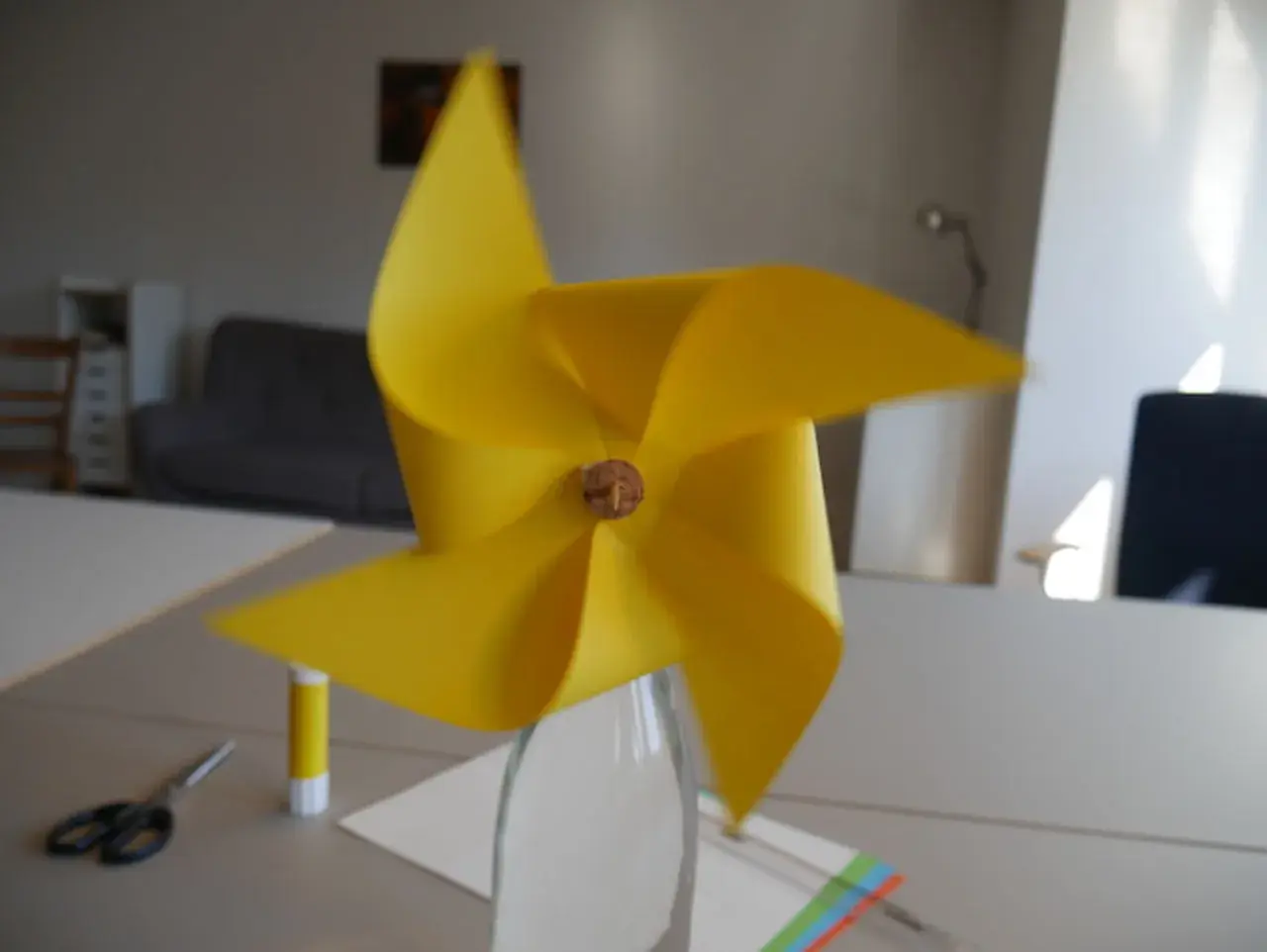 Ein gelbes Windrad, das man leicht selber bauen kann, steht in einer Glasflasche. Schere und Klebestift liegen bereit.