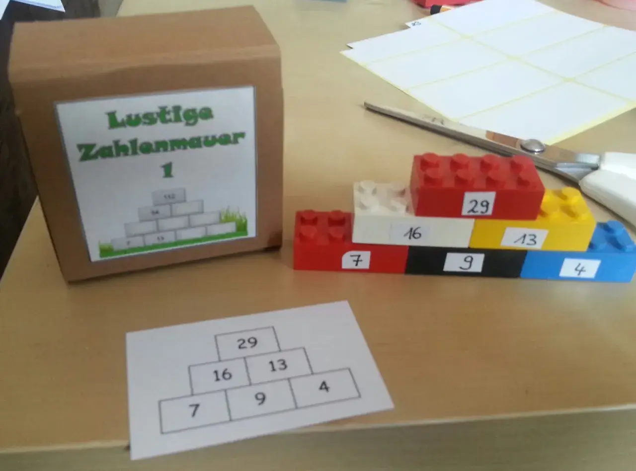 Bunte Legosteine bilden eine Zahlenmauer mit aufgeschriebenen Zahlen. Daneben eine Pappbox mit gleicher Zahlenmauer.