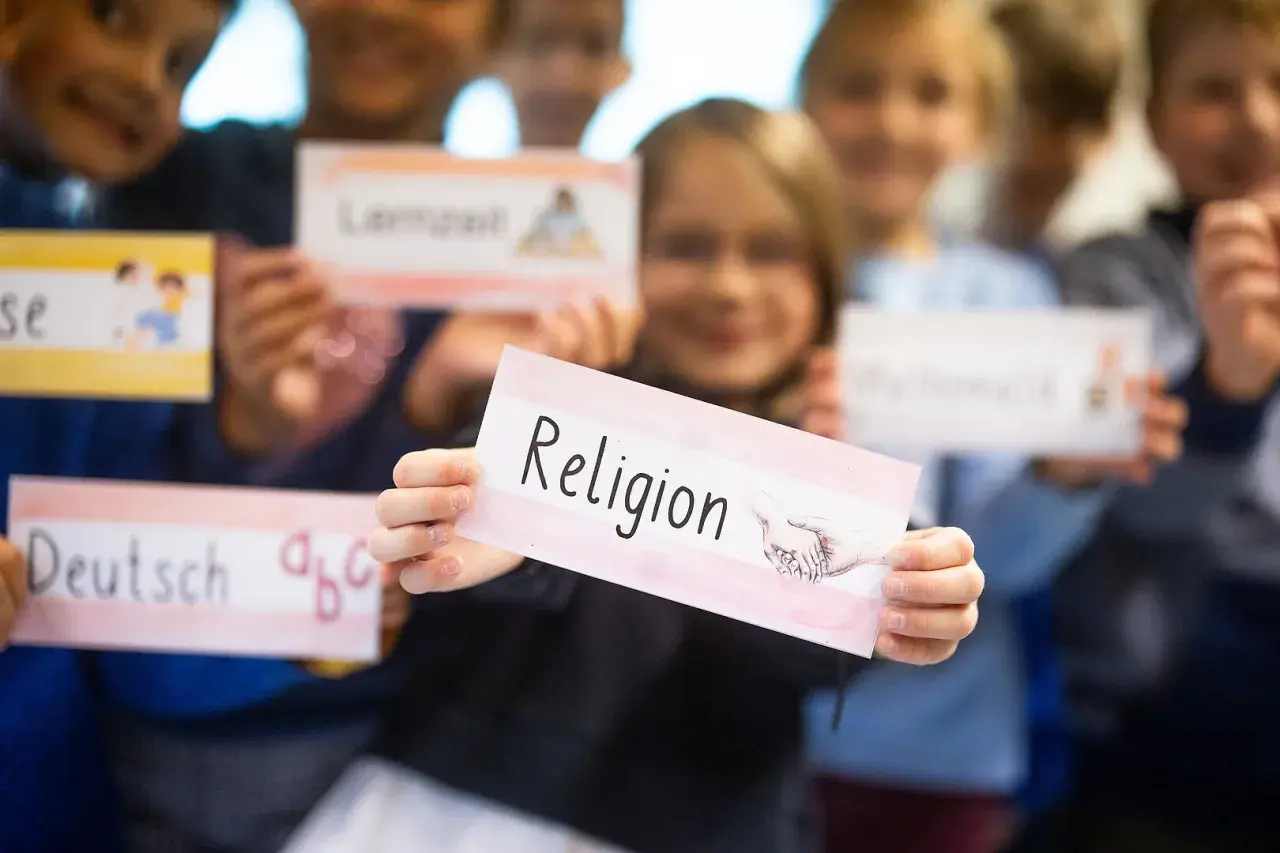Kinder der Grundschule halten Schilder hoch, eines davon mit "Religion" beschriftet. Sie lernen über verschiedene Fächer, vielleicht im Rahmen der Fastenzeit.