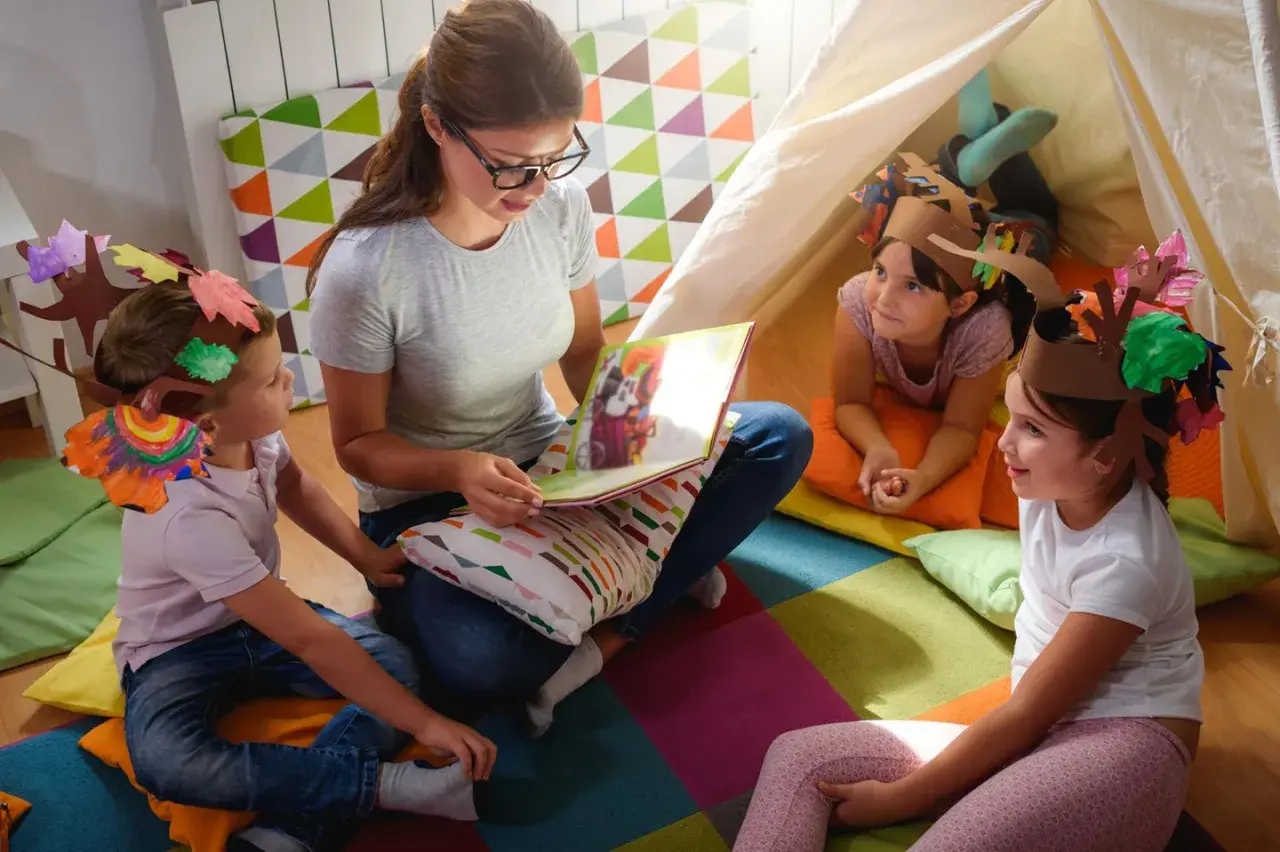 Lehrerin liest Kindern im Kindergarten eine Geschichte vor. Die Kinder tragen selbstgebastelte Baumkronen, ein Beispiel für Nachhaltigkeit im Kindergarten.
