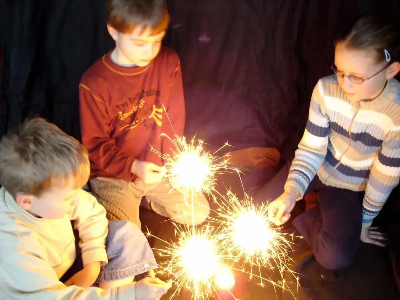 Kinder halten Wunderkerzen, die hell leuchten. Ein Junge mit rotem Pullover und ein M&auml;dchen mit Brille sind zu sehen. Ein kleiner Junge sitzt davor. Ein fr&ouml;hliches Bild, das an Feuer und Grundschule erinnert.