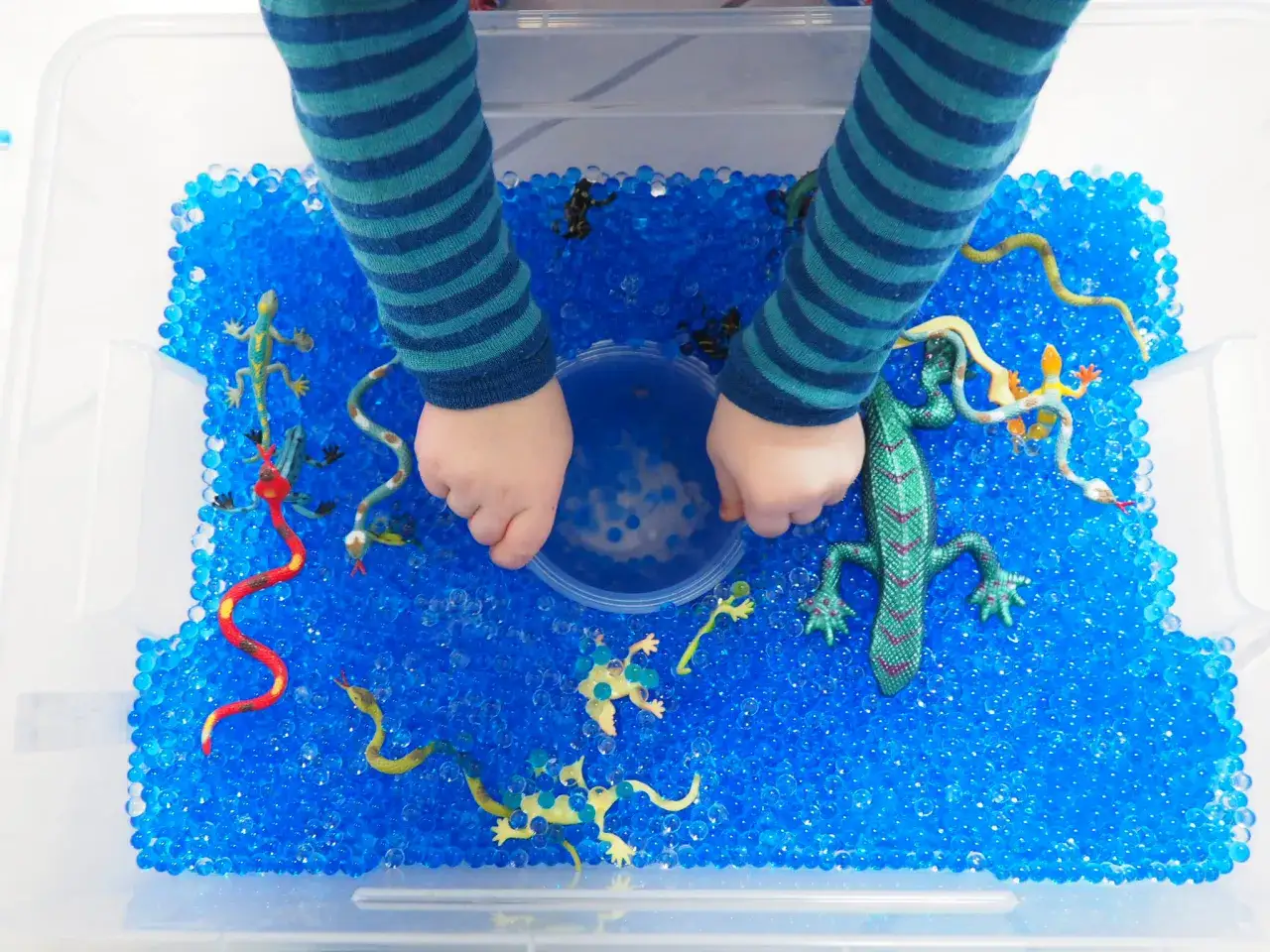 Kinderh&auml;nde erkunden eine Kiste voller blauer Wasserperlen mit Spielzeug-Echsen und Schlangen. Tolle Wasserperlen Ideen f&uuml;r sensorisches Spiel!
