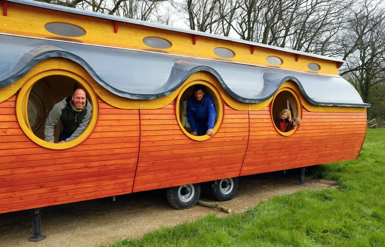 Ein bunter Bauwagen, der als Wald Kindergarten dient, mit Menschen, die aus runden Fenstern schauen.