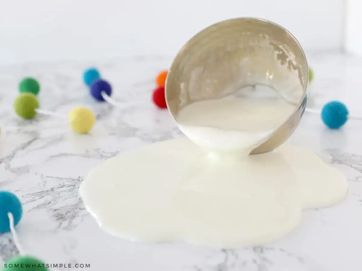 Ein umgekippter Becher mit wei&szlig;er Fl&uuml;ssigkeit, die sich auf Marmor ausbreitet. Bunte Pompons im Hintergrund deuten auf ein lustiges Oobleck-Rezept hin.
