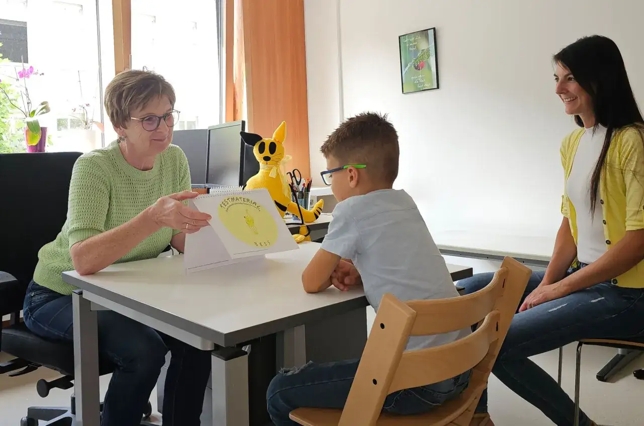 Bei der Schuleingangsuntersuchung wird spielerisch die Sprachentwicklung gepr&uuml;ft. Ein Kind schaut auf ein Heft, w&auml;hrend eine &Auml;rztin und die Mutter zusehen.
